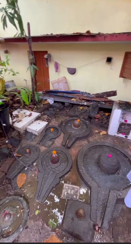 Neglected Shiva Lingams lying exposed and scattered in a courtyard, partially submerged in water and debris.