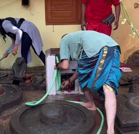 Volunteers cleaning ancient Shiva Lingams with devotion, using water hoses and brushes amid a muddy, neglected site.