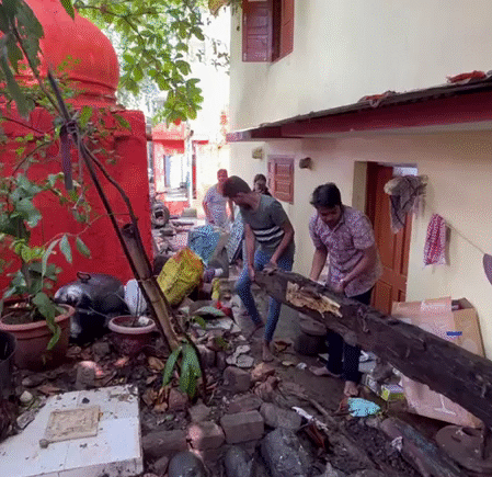 Volunteers lifting a decayed wooden beam while clearing debris near ancient Shiva Lingams under a large tree canopy.