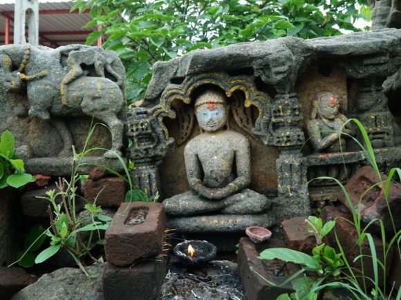 Close-up of an ancient stone carving of a meditating Jain Tirthankara, surrounded by smaller carvings, plants, and offerings with a lit oil lamp.