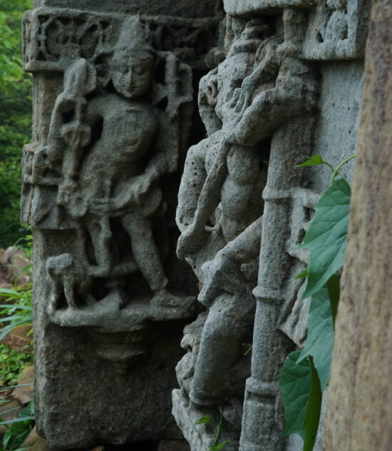 Ancient weathered stone carvings depicting human figures on a temple wall, partially covered with green leaves.