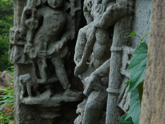 Ancient weathered stone carvings depicting human figures on a temple wall, partially covered with green leaves.